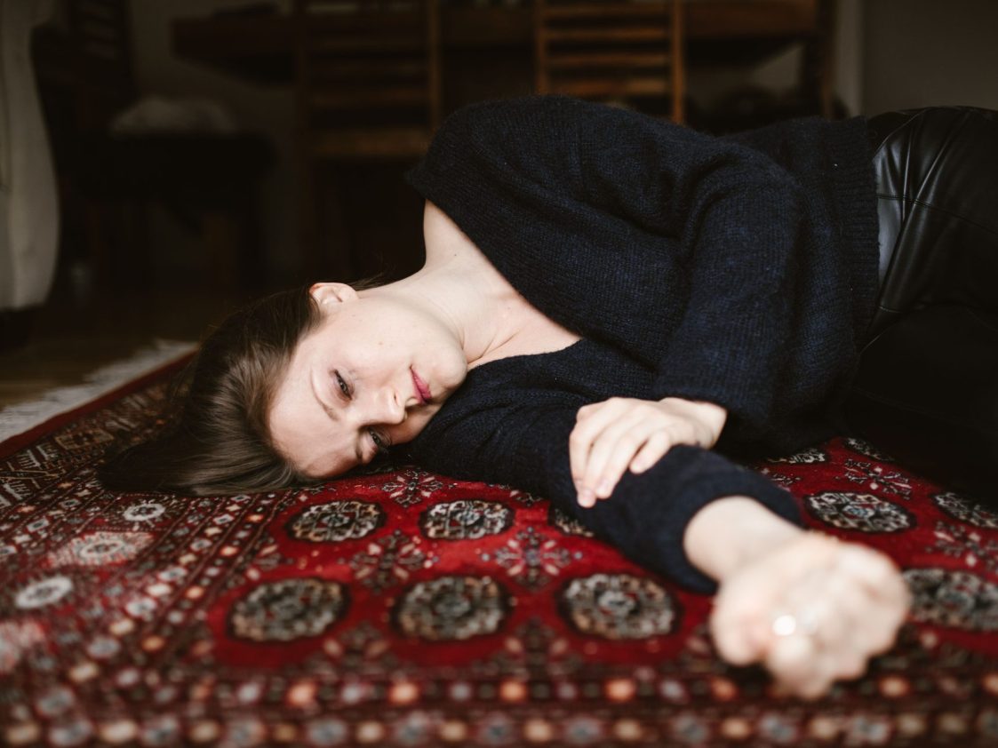 Woman lies on carpet, defeated. She has a sad, far away look in her eye and tear streaks of mascara on her face.