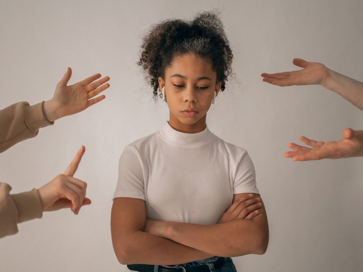 Woman stands crossed armed, looking disappointed as hands point to her disagreeably.