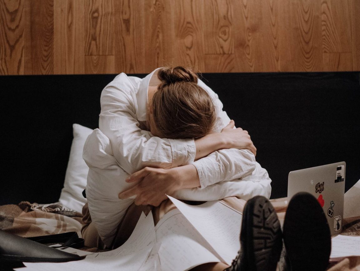 Woman sits with her head in her arms on a bed surrounded by papers, completely stressed.